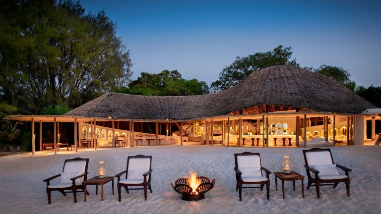 Thatched main guest area of &Beyond Mnemba Island lit at dusk with fire pit and beach chairs