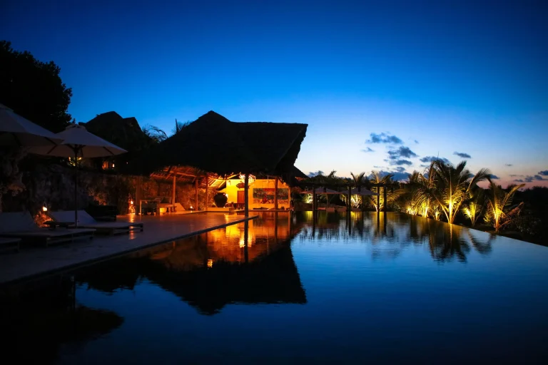 Pool and thatched buildings lit at dusk at Qambani